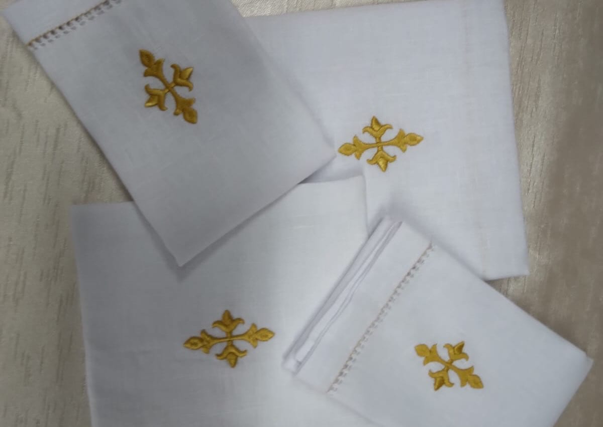 Sanguinhos de linho branco dobrados, cada um com uma cruz bordada em fio dourado.